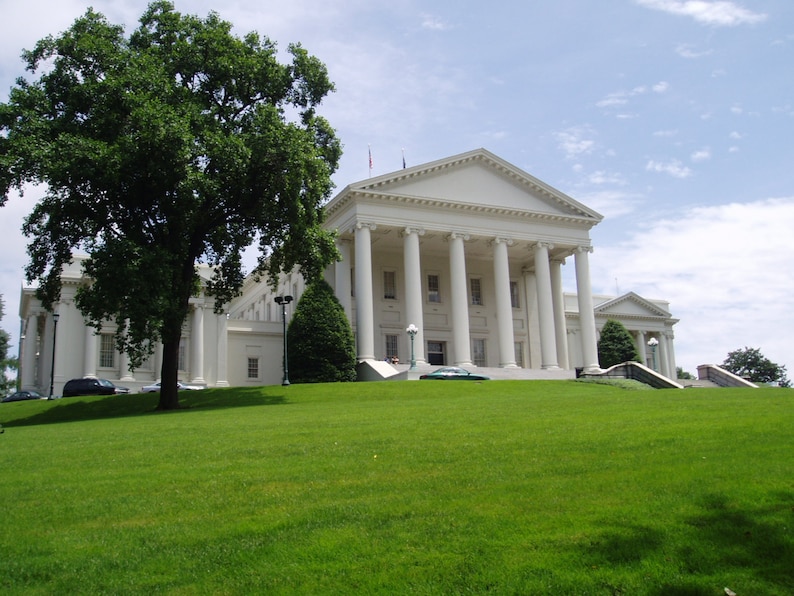 May include: A large white neoclassical building with a prominent triangular pediment supported by tall columns. The structure features multiple rectangular windows and is situated behind a wide, vibrant green lawn with a large tree on the left side.