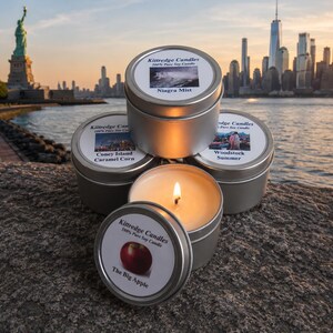 May include: A collection of silver tin candles with labels featuring images and text. One candle is lit, revealing a flame. The labels read "Niagara Mist", "Coney Island Caramel Corn", "Woodstock Summer", and "The Big Apple". The Statue of Liberty and a city skyline are in the background.