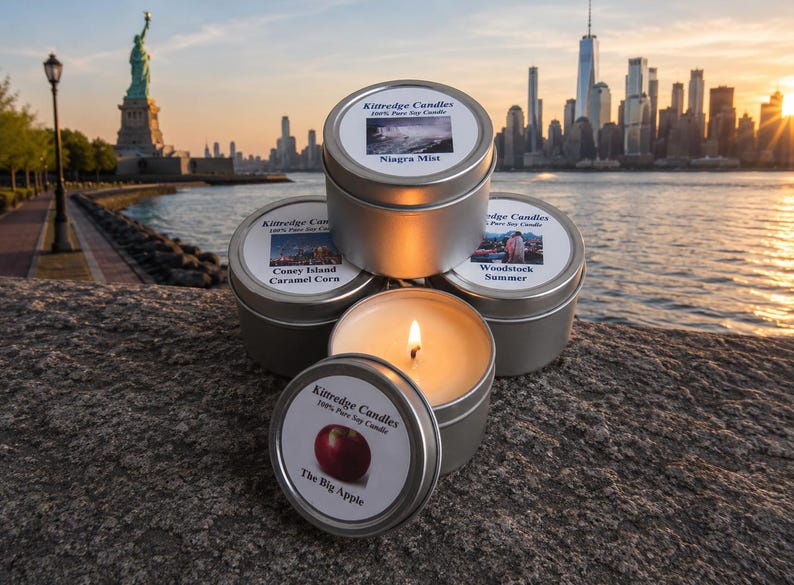 May include: A collection of silver tin candles with labels featuring images and text. One candle is lit, revealing a flame. The labels read "Niagara Mist", "Coney Island Caramel Corn", "Woodstock Summer", and "The Big Apple". The Statue of Liberty and a city skyline are in the background.