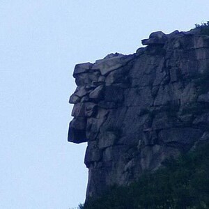 May include: A large, dark gray rock formation featuring a natural profile that resembles a human face in silhouette. The craggy cliffside displays jagged stone layers and deep crevices against a pale, light blue sky, with green foliage visible.