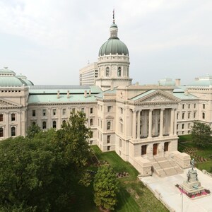 May include: An aerial view of a large, light-colored building with a prominent dome and multiple levels. The building has classical architectural features, including columns and a grand staircase. Green trees and a cloudy sky are in the background.