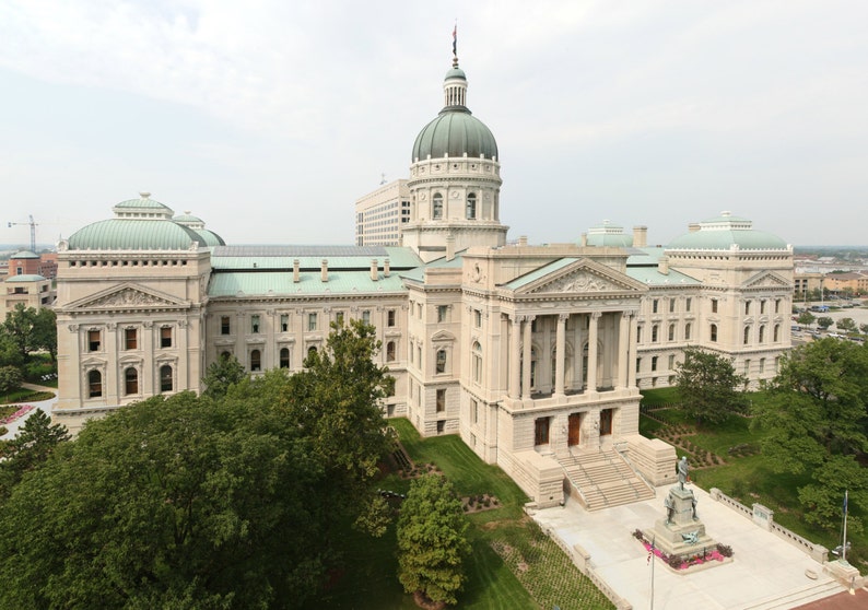 May include: An aerial view of a large, light-colored building with a prominent dome and multiple levels. The building has classical architectural features, including columns and a grand staircase. Green trees and a cloudy sky are in the background.