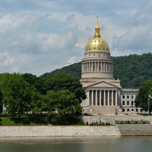 May include: The West Virginia State Capitol building features a prominent gold-leaf dome atop a neoclassical structure with white stone columns and a grand entrance, surrounded by lush green trees under a sky with scattered white clouds.