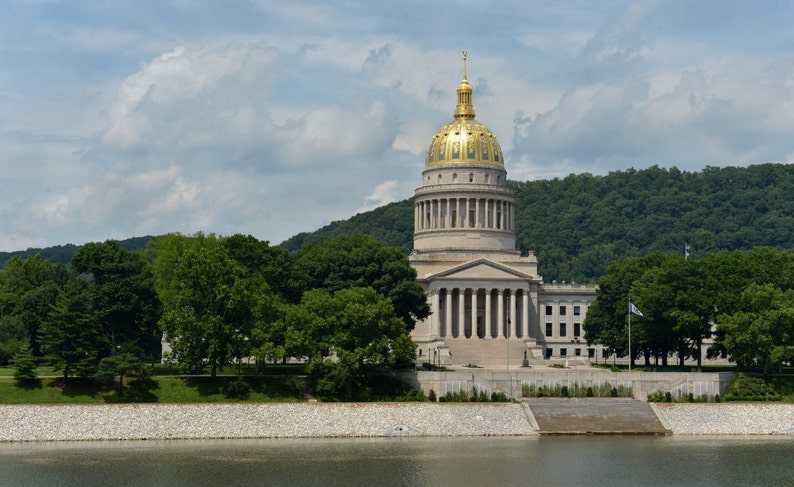 May include: The West Virginia State Capitol building features a prominent gold-leaf dome atop a neoclassical structure with white stone columns and a grand entrance, surrounded by lush green trees under a sky with scattered white clouds.