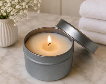 May include: A lit candle in a round, matte gray metal tin with its matching lid resting partially open on the side. The candle features a single burning wick centered in smooth, off-white wax, creating a warm glow within the container.