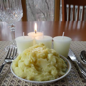 May include: A close-up of a white ceramic bowl filled with mashed potatoes. Three white candles are lit on a table with a glass of water and a place setting.