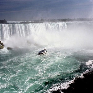 May include: A scenic view of Niagara Falls, showcasing the cascading water and the mist it creates. A boat navigates the turquoise water below the falls, with a dark, cloudy sky overhead.