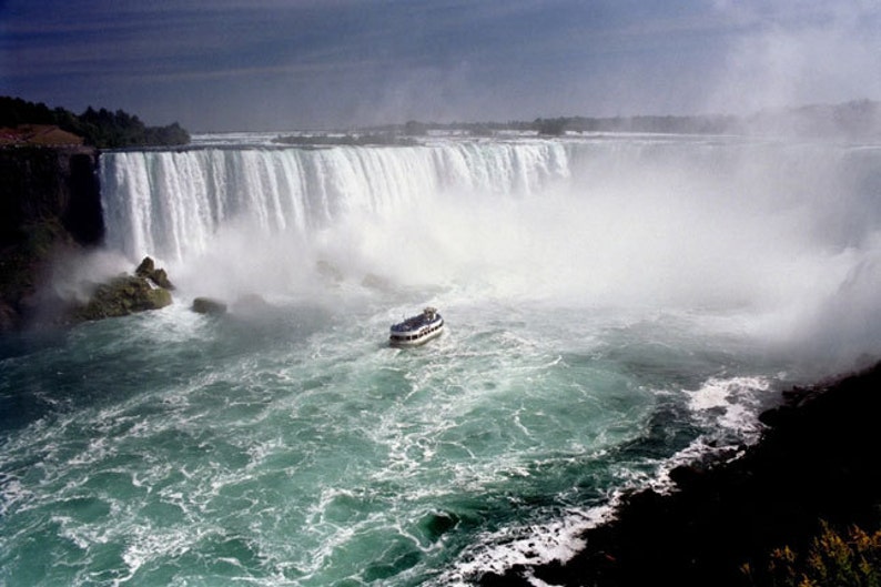 May include: A scenic view of Niagara Falls, showcasing the cascading water and the mist it creates. A boat navigates the turquoise water below the falls, with a dark, cloudy sky overhead.