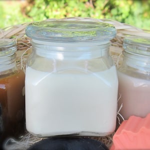 May include: Three glass jars with lids, filled with white, brown, and beige scented candles. The jars are sitting on a woven basket.