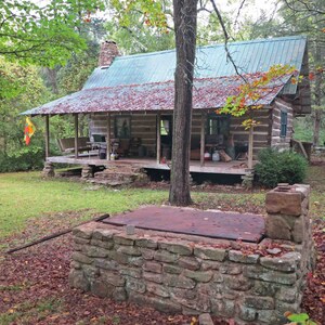 May include: A rustic log cabin with a green metal roof and a covered porch, featuring a stone chimney and a stone-walled cellar entrance in the foreground. The cabin is surrounded by trees with autumn leaves scattered on the ground and porch roof.