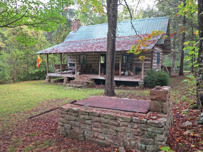 May include: A rustic log cabin with a green metal roof and a covered porch, featuring a stone chimney and a stone-walled cellar entrance in the foreground. The cabin is surrounded by trees with autumn leaves scattered on the ground and porch roof.