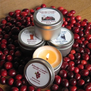 May include: Four small silver tins with labels and lids. The labels have images and text. The text on the labels reads "Kittredge Candles 100% Pure Soy Candle", "Wisconsin Dairy Farm", "Milwaukee Brewery", "Apostle Islands Mist", and "Wisconsin Cranberries". The tins are surrounded by red cranberries.
