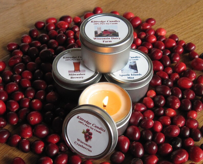 May include: Four small silver tins with labels and lids. The labels have images and text. The text on the labels reads "Kittredge Candles 100% Pure Soy Candle", "Wisconsin Dairy Farm", "Milwaukee Brewery", "Apostle Islands Mist", and "Wisconsin Cranberries". The tins are surrounded by red cranberries.