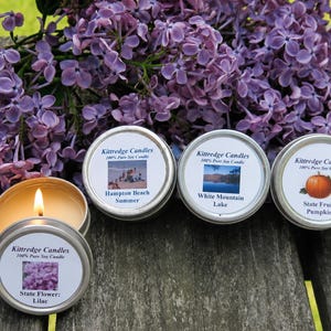 May include: Four round metal tins containing Kittredge Candles 100% Pure Soy Candle. One candle is lit, revealing yellow wax. Labels feature images of Hampton Beach, White Mountain Lake, a pumpkin, and lilacs with text identifying each scent.