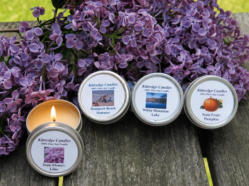 May include: Four round metal tins containing Kittredge Candles 100% Pure Soy Candle. One candle is lit, revealing yellow wax. Labels feature images of Hampton Beach, White Mountain Lake, a pumpkin, and lilacs with text identifying each scent.