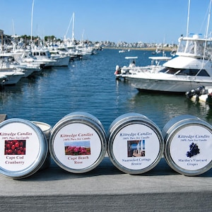 May include: Four round silver metal tins of Kittredge Candles 100% Pure Soy Candle. The labels feature images of cranberries, beach roses, a cottage, and grapes with the text Cape Cod Cranberry, Cape Beach Rose, Nantucket Cottage Breeze, and Martha's Vineyard Grapes.