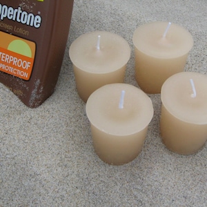 May include: Four unscented votive candles in a light beige color, sitting on a sandy surface. A bottle of Coppertone sunscreen lotion is visible in the background.