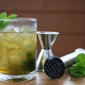 May include: A refreshing cocktail in a clear glass, garnished with mint sprigs and ice. Stainless steel jigger and muddler are next to the drink. The drink has a light green hue, suggesting a mint-based beverage.