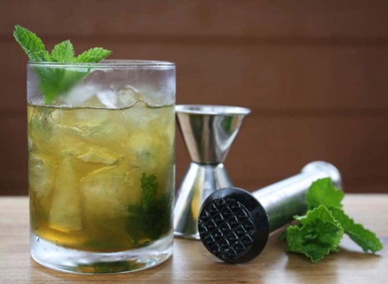 May include: A refreshing cocktail in a clear glass, garnished with mint sprigs and ice. Stainless steel jigger and muddler are next to the drink. The drink has a light green hue, suggesting a mint-based beverage.