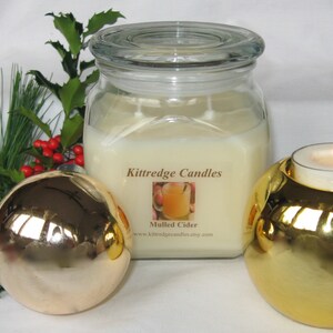 May include: A large, square glass jar candle labeled "Mulled Cider" with a lit tea light candle in a gold holder. A gold Christmas ornament and greenery with red berries are in the foreground. The candle is from Kittredge Candles.