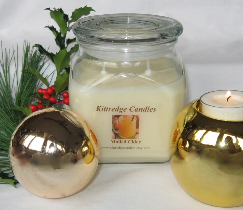 May include: A large, square glass jar candle labeled "Mulled Cider" with a lit tea light candle in a gold holder. A gold Christmas ornament and greenery with red berries are in the foreground. The candle is from Kittredge Candles.