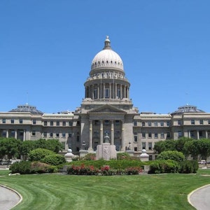 Pu&ograve; includere: Il Campidoglio dello Stato dell'Idaho &egrave; un grande edificio in pietra di colore chiaro caratterizzato da una cupola centrale prominente, una facciata simmetrica con colonne classiche e un prato anteriore con cespugli verdi e fiori rossi.