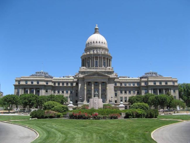 Pu&ograve; includere: Il Campidoglio dello Stato dell'Idaho &egrave; un grande edificio in pietra di colore chiaro caratterizzato da una cupola centrale prominente, una facciata simmetrica con colonne classiche e un prato anteriore con cespugli verdi e fiori rossi.