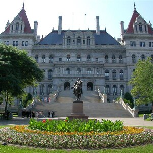 May include: A large, ornate building with a light gray facade, red roof accents, and multiple windows. A statue of a man on a horse stands in front of a grand staircase. A flower bed with colorful flowers is in the foreground.