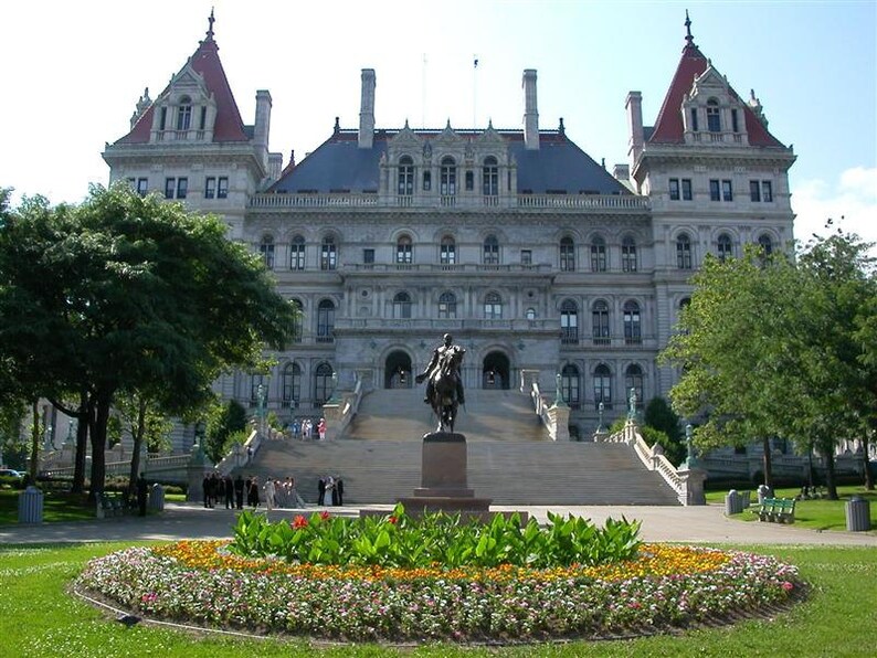 May include: A large, ornate building with a light gray facade, red roof accents, and multiple windows. A statue of a man on a horse stands in front of a grand staircase. A flower bed with colorful flowers is in the foreground.