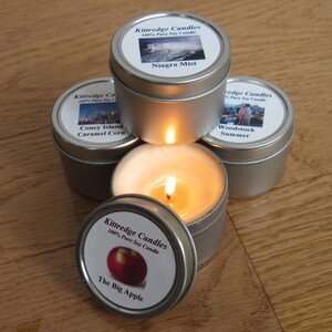 May include: A collection of small, silver-colored tin candles with labels featuring scenic images and text. One candle is lit, with a bright flame. The labels read "Niagara Mist", "Coney Island Caramel Corn", "Woodstock Summer", and "The Big Apple".