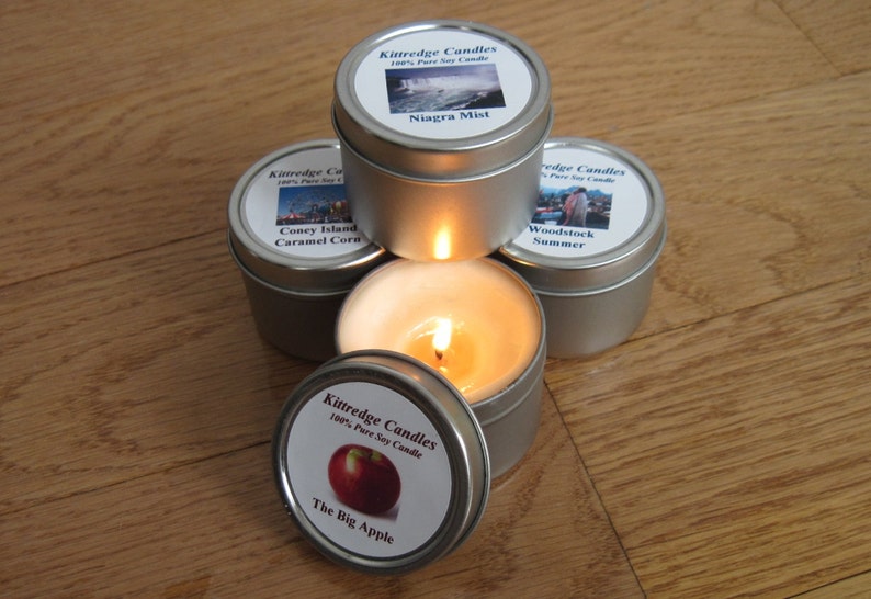 May include: A collection of small, silver-colored tin candles with labels featuring scenic images and text. One candle is lit, with a bright flame. The labels read "Niagara Mist", "Coney Island Caramel Corn", "Woodstock Summer", and "The Big Apple".