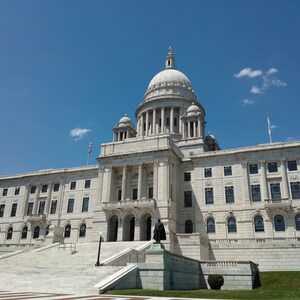 May include: A large, light-colored building with a prominent dome and multiple columns, set against a bright blue sky. The building has many windows and is approached by a wide set of stairs. Flags are visible on the building.