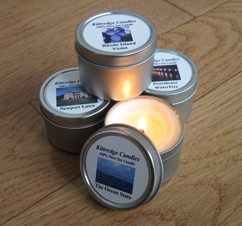 May include: A collection of small, silver-colored tin candles with labels featuring images and text. One candle is lit, showing a warm, yellow flame. The labels read "Kittredge Candles" and include phrases like "Rhode Island Violet" and "The Ocean State."
