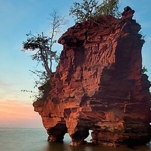 May include: A large, red sandstone rock formation with a small tree growing on top. The rock is partially submerged in the water, creating a natural archway. The sky is a soft blue with pink and orange hues.