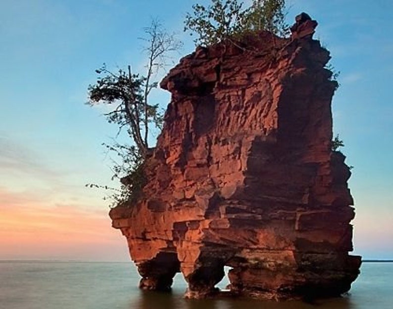 May include: A large, red sandstone rock formation with a small tree growing on top. The rock is partially submerged in the water, creating a natural archway. The sky is a soft blue with pink and orange hues.