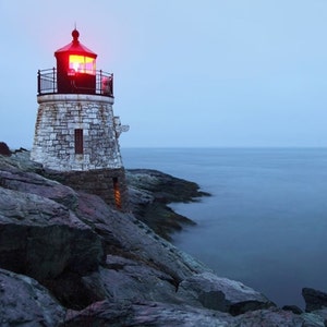 May include: A stone lighthouse with a red light on top, perched on a rocky cliff overlooking the ocean. The sky is a muted blue, and the water is calm. The lighthouse is made of light-colored stone.