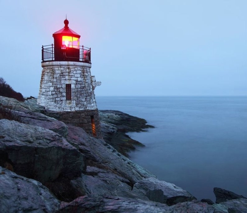 May include: A stone lighthouse with a red light on top, perched on a rocky cliff overlooking the ocean. The sky is a muted blue, and the water is calm. The lighthouse is made of light-colored stone.