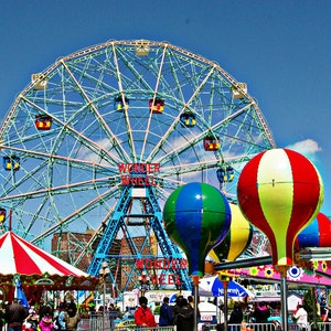 May include: A vibrant image of Coney Island's Wonder Wheel and amusement park rides under a bright blue sky. The Ferris wheel is the focal point, with colorful gondolas and the words "Wonder Wheel" displayed. Balloon-shaped ride cars add to the festive atmosphere.