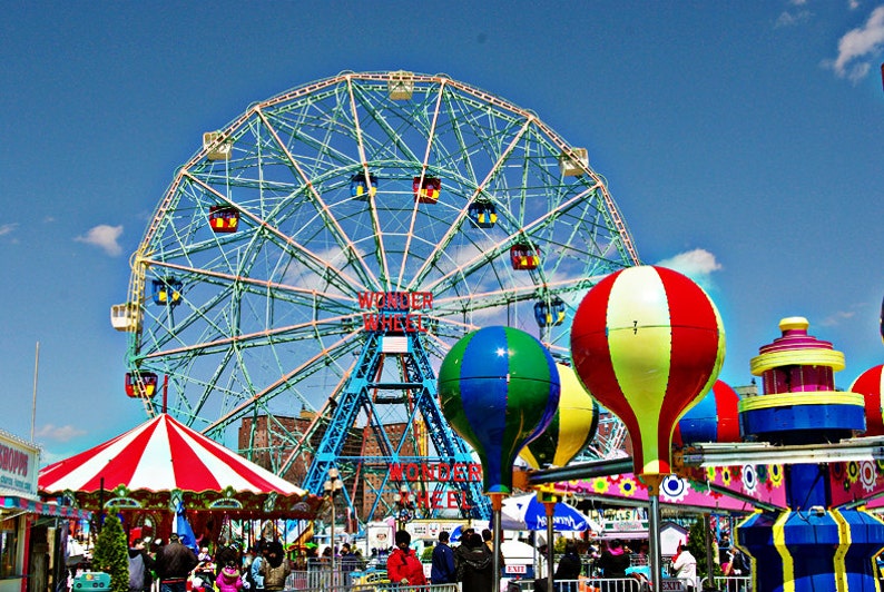 May include: A vibrant image of Coney Island's Wonder Wheel and amusement park rides under a bright blue sky. The Ferris wheel is the focal point, with colorful gondolas and the words "Wonder Wheel" displayed. Balloon-shaped ride cars add to the festive atmosphere.