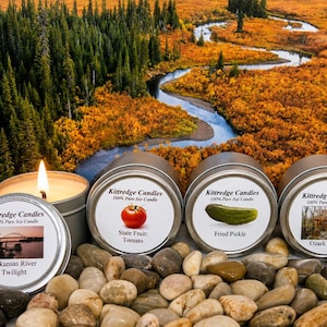 May include: Four round metal tins of Kittredge Candles 100% Pure Soy Candle. One tin is open with a lit wick. Labels feature images of a bridge, a tomato, a pickle, and a cabin with text: Arkansas River Twilight, State Fruit: Tomato, Fried Pickle, Ozark Cabin.