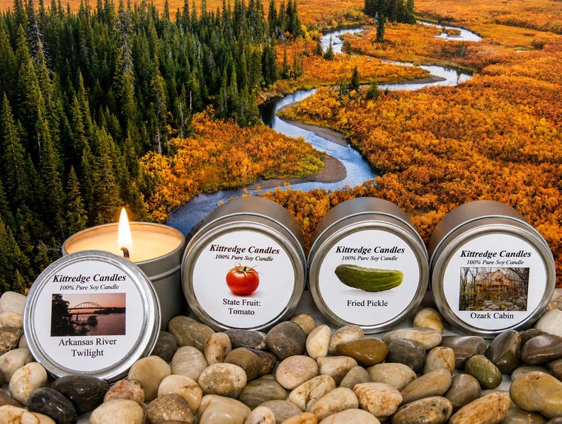 May include: Four round metal tins of Kittredge Candles 100% Pure Soy Candle. One tin is open with a lit wick. Labels feature images of a bridge, a tomato, a pickle, and a cabin with text: Arkansas River Twilight, State Fruit: Tomato, Fried Pickle, Ozark Cabin.