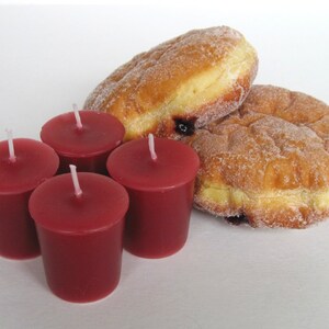 May include: An image featuring four small, burgundy-colored votive candles with white wicks, alongside two sugared donuts. The donuts are golden brown with a visible filling. The items are arranged on a white surface.