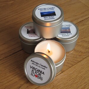May include: Four round silver metal tins containing soy candles. Three tins are closed with labels reading Kittredge Candles, Blue Ridge Mountains, Arlington Cherry, and Virginia Beach Summer. One open tin features a lit candle and a lid with the text Virginia Is For Lovers.