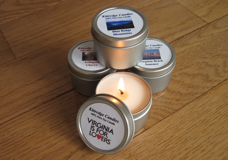 May include: Four round silver metal tins containing soy candles. Three tins are closed with labels reading Kittredge Candles, Blue Ridge Mountains, Arlington Cherry, and Virginia Beach Summer. One open tin features a lit candle and a lid with the text Virginia Is For Lovers.