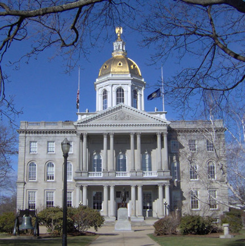 May include: The New Hampshire State House is a gray stone building featuring a prominent gold-colored dome topped with a golden eagle statue. The structure has a classical facade with multiple columns, symmetrical windows, and a central entrance.