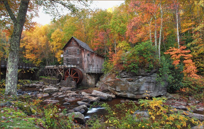 May include: A rustic wooden grist mill with a large red water wheel sits beside a rocky stream. The structure features multiple windows and a shingled roof, surrounded by a dense forest displaying vibrant autumn foliage in shades of orange, yellow, and red.