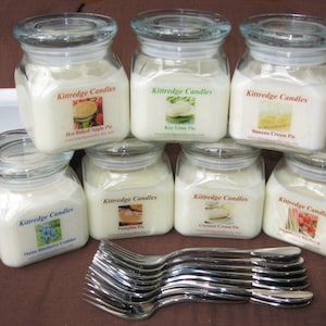 May include: Eight glass jars with white candles and labels that say "Kittredge Candles" and the name of a pie flavor. The jars are arranged in two rows of four. The jars are on a brown surface with a stack of white plates in the background.