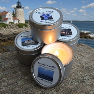 May include: Several small, silver-colored candle tins are stacked on a wooden surface. One tin is open, with a lit candle inside. The labels read "Kittredge Candles" and feature images and text like "Rhode Island Violet" and "The Ocean State."