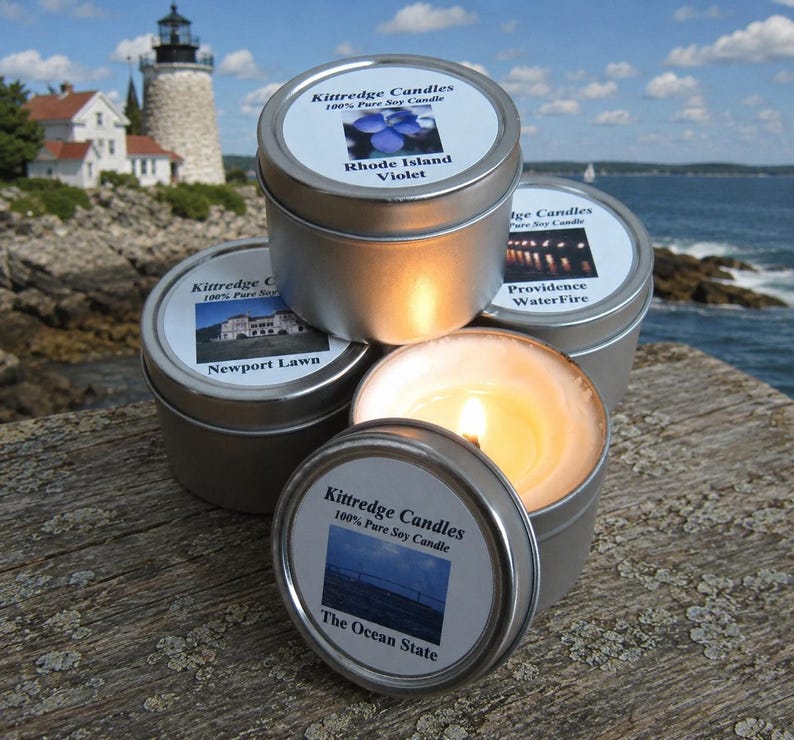 May include: Several small, silver-colored candle tins are stacked on a wooden surface. One tin is open, with a lit candle inside. The labels read "Kittredge Candles" and feature images and text like "Rhode Island Violet" and "The Ocean State."