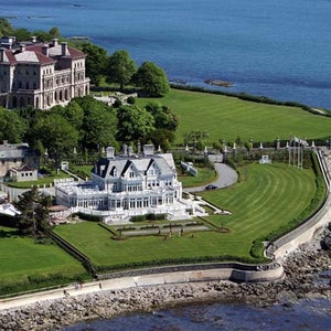 May include: Aerial view of a grand estate with multiple buildings, lush green lawns, and a coastal setting. The main structures are large, with intricate architectural details. The property overlooks the ocean, with a stone seawall and rocky shoreline.